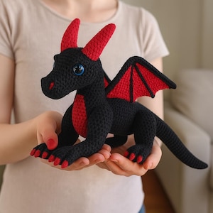 May include: A handmade black crochet dragon with red accents. The dragon features red horns, wings, and chest markings. It has blue eyes and is held in someone's hands.