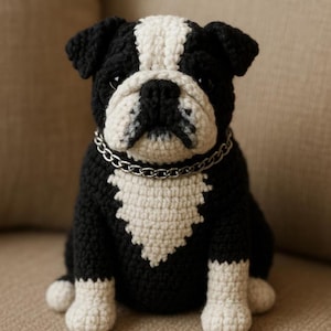 Puede incluir: Un bulldog de ganchillo hecho a mano en blanco y negro, con un collar de cadena plateada. El perro está sentado, mostrando sus distintivas marcas. Un artículo encantador y artesanal.