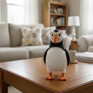 May include: A handmade crochet puffin bird with a white belly, black back and wings, and an orange beak and feet. The bird has white fluffy hair and is standing on a wooden table. The puffin is a decorative item.