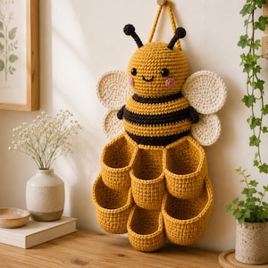 Puede incluir: Un colgante de pared de ganchillo con forma de abeja, cuerpo a rayas amarillas y negras, dos alas blancas y seis pequeños bolsillos amarillos dispuestos en forma de panal. La abeja tiene antenas negras, ojos negros y mejillas rosadas.