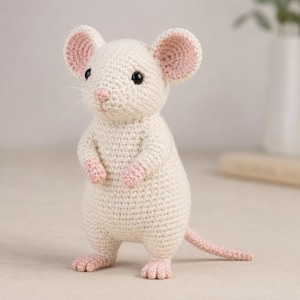 Pode incluir: Uma figura de rato em crochê feita de fio creme com detalhes em rosa suave no interior das orelhas, nariz, patas e cauda. O rato apresenta olhos de contas pretas, um corpo arredondado e uma cauda longa, em posição vertical.