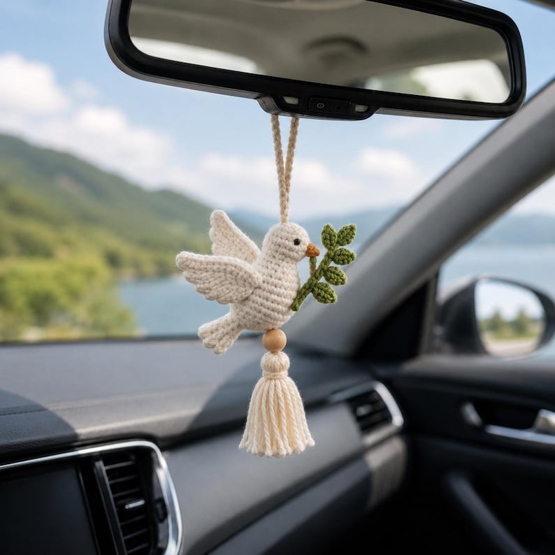 May include: A crocheted white dove car charm, holding a green olive branch in its beak. The dove hangs from a string, with a wooden bead and tassel. The charm is suspended from a car's rearview mirror, with a scenic background.