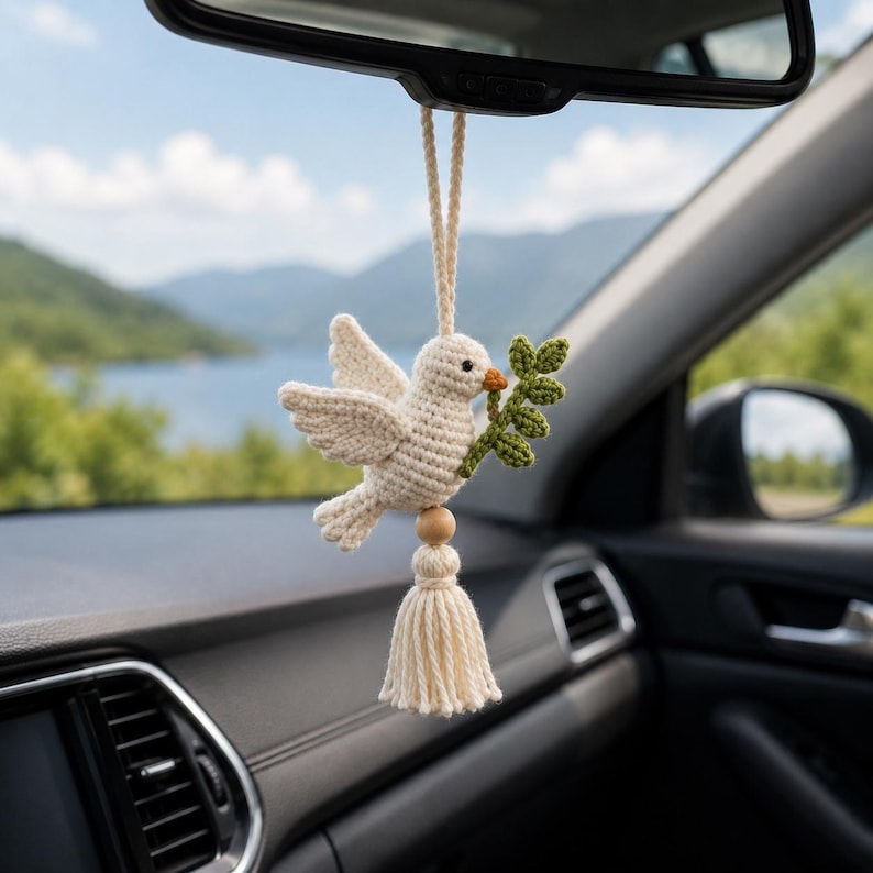 May include: A crocheted dove car charm, cream-colored with a green olive branch in its beak, hangs from a car's rearview mirror. A wooden bead and tassel complete the design. The background shows a lake and mountains.