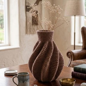Puede incluir: Un jarrón marrón tejido a ganchillo con una textura y un diseño retorcido único. El jarrón contiene flores secas y está sobre una mesa de madera. Una taza, un cuenco pequeño y libros también están sobre la mesa. La habitación tiene un ambiente cálido.