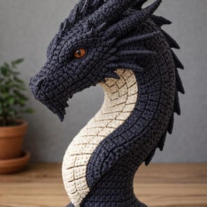 Op de afbeelding: Gehaakte drakenhoofdsculptuur in donkerblauw en crème. De draak heeft gedetailleerde schubben, hoorns en oranje ogen. De sculptuur wordt tentoongesteld op een houten oppervlak. De nek van de draak heeft een crèmekleurig patroon.