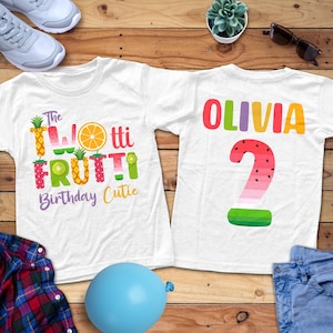 May include: Two white t-shirts with colorful fruit-themed designs. One shirt says "The Twotti Frutti Birthday Cutie" with fruit graphics. The other shirt has the name "Olivia" and a watermelon-themed number "2".