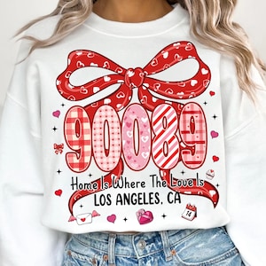 May include: White sweatshirt with a large red bow design featuring hearts. The design includes the numbers "90089" in various patterns, along with the text "Home Is Where The Love Is LOS ANGELES, CA". Small heart and Valentine's Day themed graphics are scattered around the design.