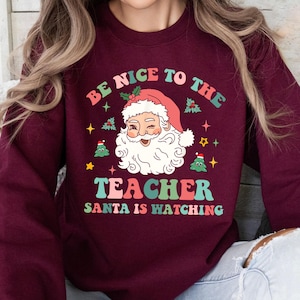 May include: A maroon sweatshirt with a retro design featuring Santa Claus winking. The text reads "BE NICE TO THE TEACHER SANTA IS WATCHING" in a colorful, playful font. Christmas trees and stars surround Santa.