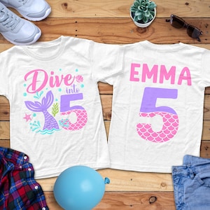 May include: Two white t-shirts with mermaid-themed graphics. One shirt says "Dive into 5" with a mermaid tail design. The other shirt has the name "EMMA" and the number "5". The graphics are in pink, purple, and teal.