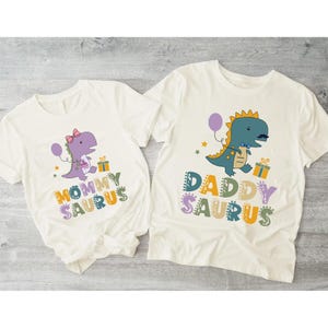 May include: Two off-white t-shirts with dinosaur graphics. One shirt says "Mommy Saurus" with a purple dinosaur wearing a bow. The other says "Daddy Saurus" with a blue dinosaur. Both have colorful text and gift box graphics.