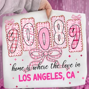 Puede incluir: Cojín blanco con elementos decorativos rosas. El cojín presenta los números "90089" en un diseño rosa y blanco, con un gran lazo en la parte inferior. También es visible el texto "home is where the love in LOS ANGELES, CA".