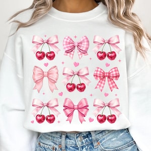 May include: White sweatshirt featuring a pattern of pink bows, some with cherries, and hearts. The bows vary in style, including solid pink and gingham patterns. Small pink hearts and stars are scattered around the bows.