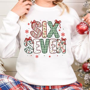 Könnte beinhalten: Weißes Sweatshirt mit den Worten "SIX SEVEN" in einem festlichen Design. Die Buchstaben sind mit roten und grünen Mustern verziert, darunter Streifen, Karos und Weihnachtsmotive. Rote Schleifen und Schneeflocken akzentuieren das Design.
