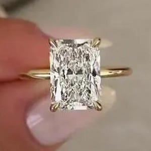 Può includere: Un anello di fidanzamento in oro con diamante taglio radiant. Il diamante rettangolare è il fulcro, con una superficie scintillante e sfaccettata. La sottile fascia in oro esalta la brillantezza del diamante. Un gioiello senza tempo.