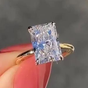 Puede incluir: Anillo de oro con un diamante de talla radiante. El diamante rectangular es el punto focal, reflejando la luz. El anillo se muestra sobre un fondo borroso, resaltando el brillo de la joya.