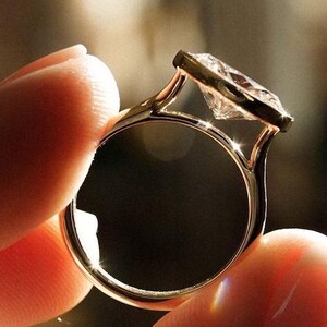 Puede incluir: Un anillo de oro con una gran gema transparente de talla cuadrada. El anillo se sostiene, mostrando el intrincado diseño y la forma en que la luz incide en la piedra. La banda es delgada y elegante, con un engaste clásico.