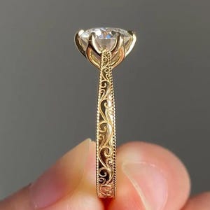 Op de afbeelding: Een close-up van een gouden ring met een grote, heldere edelsteen. De ring heeft een sierlijk, gegraveerd ontwerp op de band en rond de edelsteen setting. De ring wordt tegen een neutrale achtergrond gehouden.