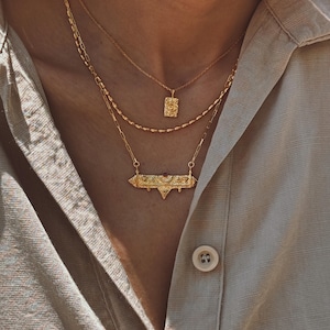 Può includere: Tre collane d'oro di diverse lunghezze. La collana superiore ha un piccolo ciondolo rettangolare. La collana centrale è una catena delicata. La collana inferiore presenta un ciondolo a barra con un design geometrico. Le collane sono a strati e indossate sopra una camicia beige.