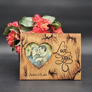 May include: Wooden picture frame with a heart-shaped photo opening. The frame has a rustic design with the words "Love You" and the names "Austin & Katie" engraved. Red flowers are in the background.