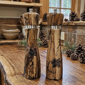 May include: Two wooden salt and pepper grinders featuring a natural wood grain with dark, branching fractal patterns and engraved pinecone designs. Each mill has a cylindrical body, a silver-colored metal ring, and a silver top adjustment knob.