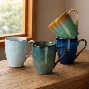 May include: Four ceramic mugs with glossy reactive glazes. One is light blue, one is dark blue, one is a deep teal, and one is a gradient of yellow to green. Each mug features a curved handle and a smooth, rounded cylindrical body shape.