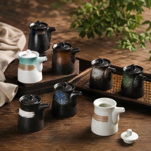 May include: A collection of eight small ceramic soy sauce dispensers with lids, featuring various textured finishes in black, white, brown, and green tones, each with a side spout and a cylindrical body, arranged on wooden surfaces.