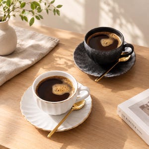 May include: Two ceramic coffee cups with matching saucers, one in matte black with a speckled finish and one in white with a ribbed texture. Both cups feature gold-toned spoons and are filled with dark coffee, presented on a wooden surface.