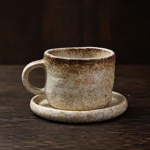 May include: A ceramic coffee cup and matching saucer featuring a speckled off-white and brown glaze. The cup has a thick, rounded handle and an uneven, textured surface. The saucer is circular with a raised edge, sharing the same speckled finish.