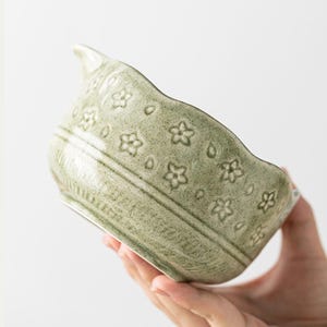 May include: A light green ceramic bowl with a spout and floral pattern. The bowl has a textured surface and is held by a hand. The floral pattern includes small flowers and teardrop shapes. The bowl is likely used for serving or decoration.