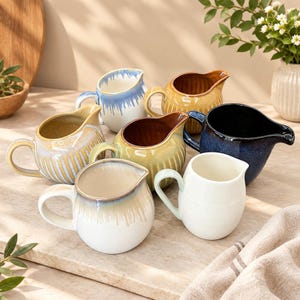 May include: A collection of six ceramic creamers in various colors including blue, yellow, brown, and white. Each creamer features a handle and a pouring spout, with some displaying ribbed textures and others showing a smooth, glazed drip finish.