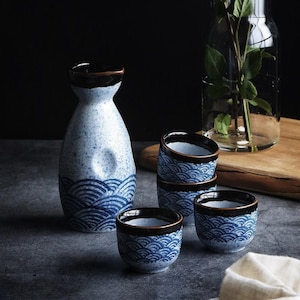 May include: A sake set featuring a blue and white ceramic carafe and matching cups. The carafe has a dark brown rim and a wave pattern. The cups also have a wave pattern and dark brown rims. A clear glass vase with greenery is in the background.