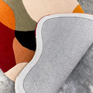 A colorful, abstract area rug with a modern design. The rug features a pattern of overlapping shapes in shades of red, orange, beige, green, and black. The underside of the rug is gray with a white border. Premium Hand Tufted Wool Rug – Modern Rust Beige Floor Rug for Bedroom & Lounge
Elegant Modern Area Rug – Handcrafted Rust Red Beige Tufted Carpet