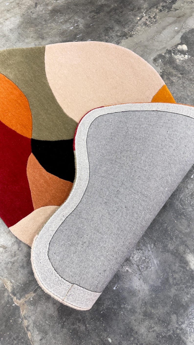 A colorful, abstract area rug with a modern design. The rug features a pattern of overlapping shapes in shades of red, orange, beige, green, and black. The underside of the rug is gray with a white border. Premium Hand Tufted Wool Rug – Modern Rust Beige Floor Rug for Bedroom & Lounge
Elegant Modern Area Rug – Handcrafted Rust Red Beige Tufted Carpet