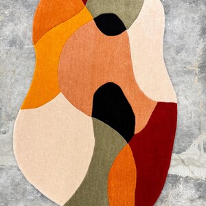Organic-shaped area rug with a modern design. The rug features a combination of colors, including orange, red, black, beige, and olive green. The rug is placed on a concrete floor. Soft Hand Tufted Wool Rug – Contemporary Rust Red Beige Living Room Rug. Designer Abstract Rug – Hand Tufted Rust Beige Carpet for Modern Homes
