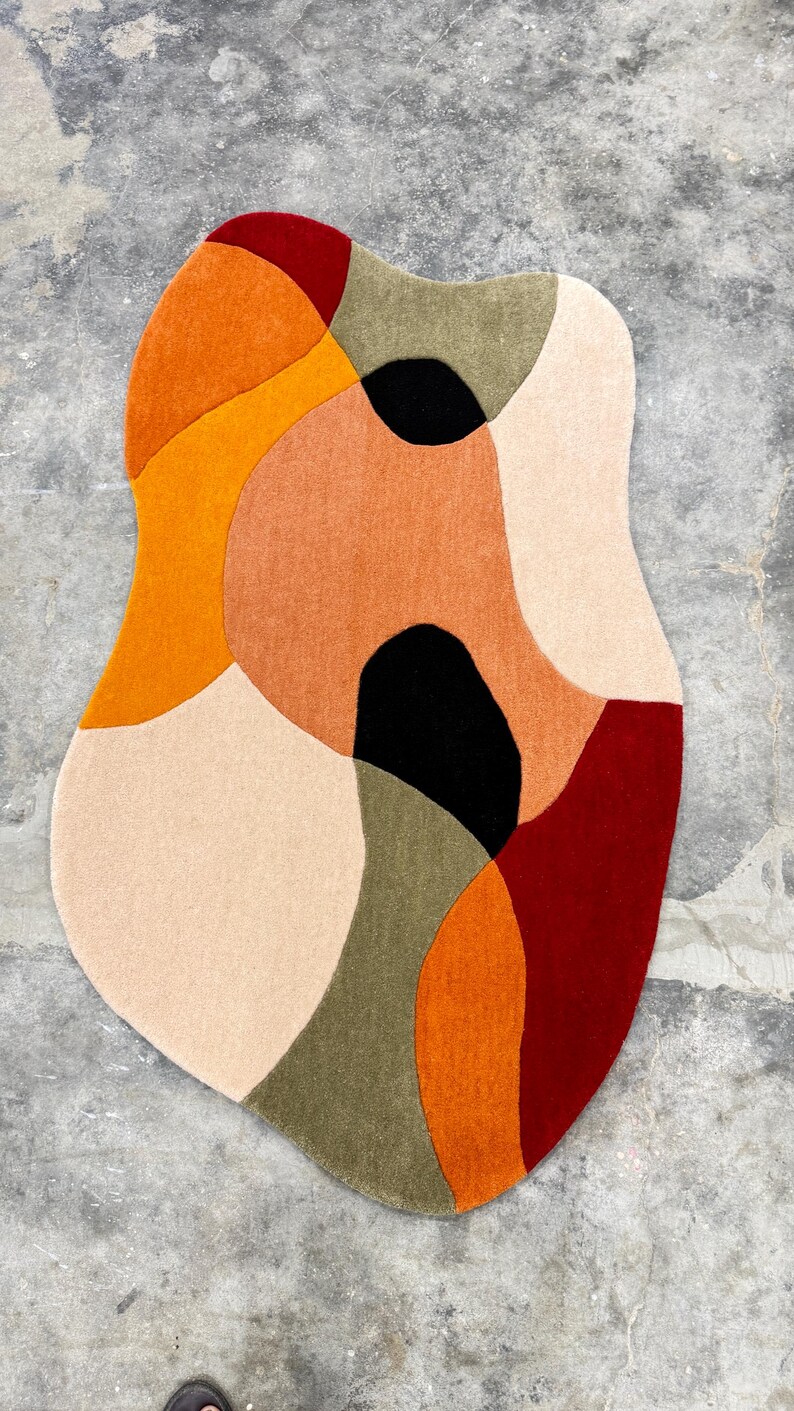 Organic-shaped area rug with a modern design. The rug features a combination of colors, including orange, red, black, beige, and olive green. The rug is placed on a concrete floor. Soft Hand Tufted Wool Rug – Contemporary Rust Red Beige Living Room Rug. Designer Abstract Rug – Hand Tufted Rust Beige Carpet for Modern Homes