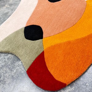 A colorful, abstract rug with a unique shape. The rug features a combination of colors, including cream, peach, olive green, black, red, and orange. The rug's design is modern and artistic, suitable for a living room or bedroom.