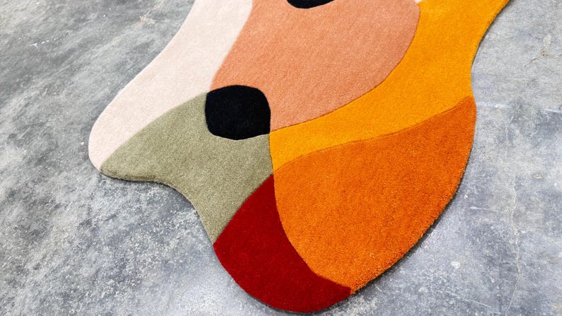 A colorful, abstract rug with a unique shape. The rug features a combination of colors, including cream, peach, olive green, black, red, and orange. The rug's design is modern and artistic, suitable for a living room or bedroom.