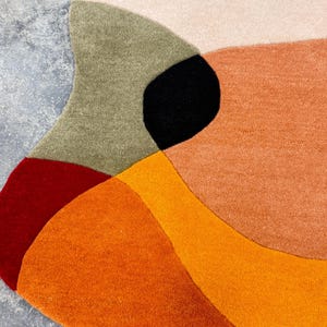 A colorful, abstract rug with a modern design. The rug features a combination of warm colors, including shades of orange, red, and beige, with a black accent. The rug's irregular shape adds to its unique aesthetic.