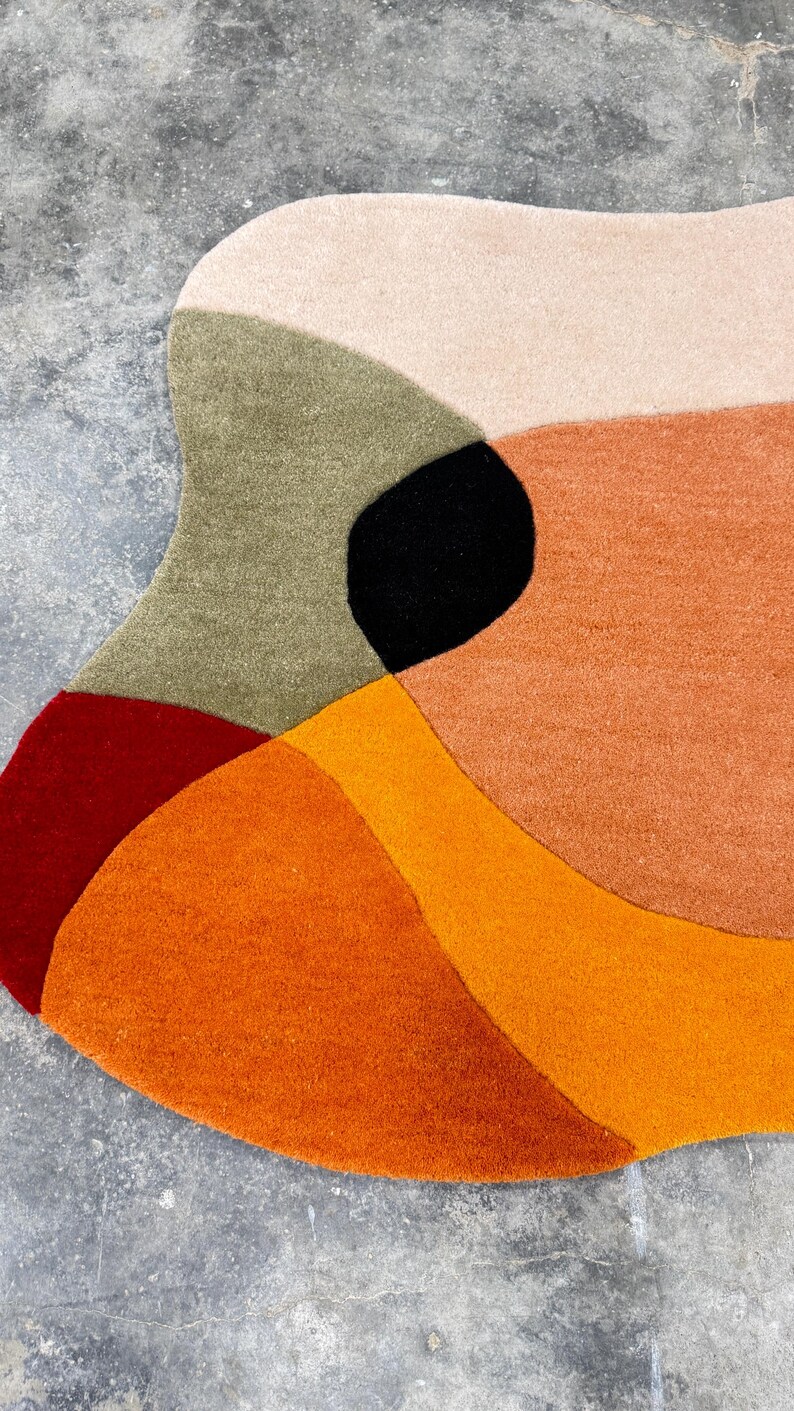 A colorful, abstract rug with a modern design. The rug features a combination of warm colors, including shades of orange, red, and beige, with a black accent. The rug's irregular shape adds to its unique aesthetic.