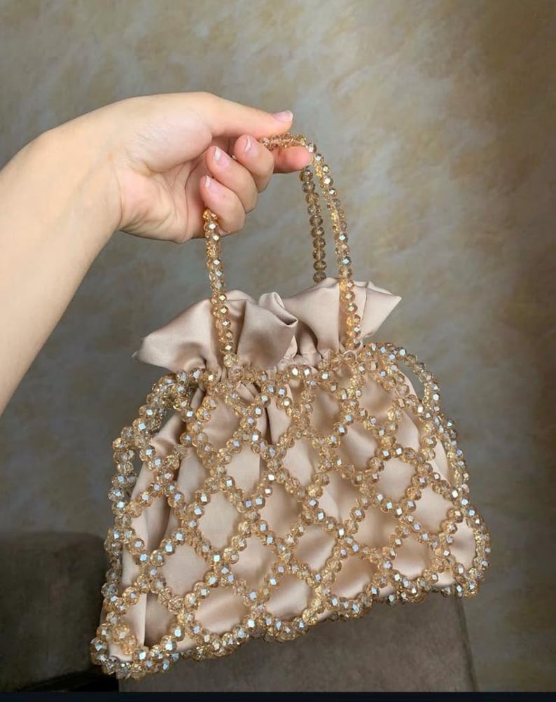 May include: A beige satin drawstring bag with a beaded overlay. The bag features a grid-like pattern of clear, faceted beads. The handles are also made of beaded strands. The bag is held by a hand.