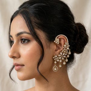 May include: An ornate gold-tone ear cuff featuring a curved design that follows the contour of the ear. The piece is embellished with numerous small white pearls and clear crystals, with several teardrop-shaped pearl dangles hanging from the base.