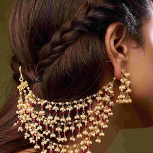 May include: A gold-coloured hair accessory and matching earrings. The jewellery features cascading strands of small, white beads interspersed with red beads. The hair is styled in a braid. The background is a solid, muted green.