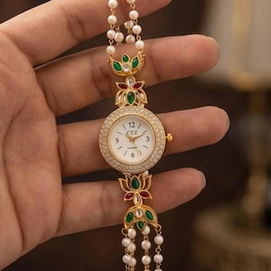 May include: Gold-toned watch with a white face, adorned with small diamonds around the bezel. The watch features a pearl and gold chain strap, and decorative green and red accents. The watch face displays the text "CTZ" and the numbers 12, 3, 6, and 9.
