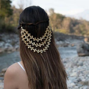 May include: A gold-colored hair accessory with a floral design. The accessory is worn in the hair and cascades down the back. The hair is long and brown. The background is a natural outdoor setting with a river.