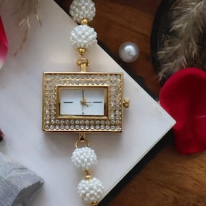 May include: A gold-coloured wristwatch with a rectangular face, adorned with small crystals. The watch strap features white beaded spheres connected by gold-coloured links. The watch is set against a neutral background with decorative elements.