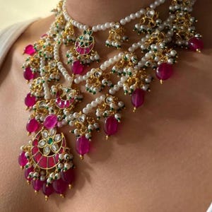 May include: A multi-strand necklace featuring white pearls, gold-toned accents, and vibrant fuchsia-colored gemstones. The necklace has a layered design with intricate detailing, including small green beads and ornate pendants.