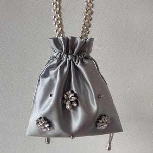 May include: A silver satin drawstring bag with a beaded handle. The bag is decorated with sparkling crystal floral accents and small silver studs. The drawstring closure has silver tassels.