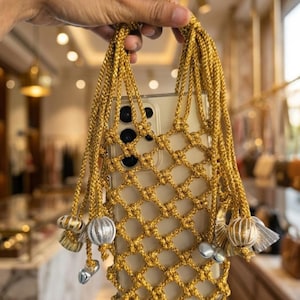 May include: A gold macrame phone bag with a clear phone case inside. The bag has a woven net design and decorative tassels with silver and gold beads. The bag is held by a hand.