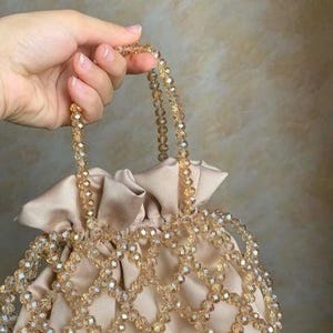 May include: A beige satin handbag with a beaded overlay and handles. The bag features a drawstring closure and is adorned with clear, faceted beads. The handles are also made of the same beaded material, creating a cohesive and elegant design.