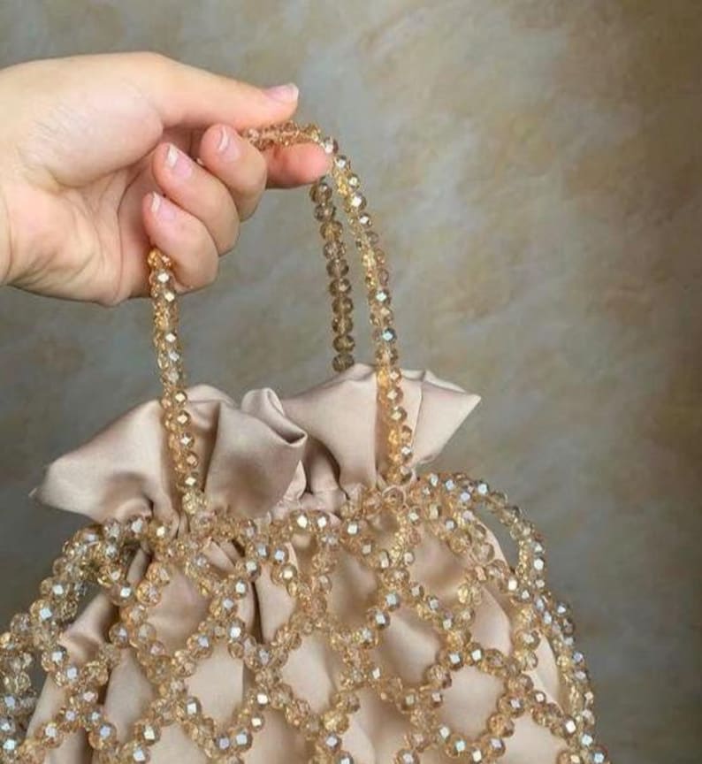 May include: A beige satin handbag with a beaded overlay and handles. The bag features a drawstring closure and is adorned with clear, faceted beads. The handles are also made of the same beaded material, creating a cohesive and elegant design.